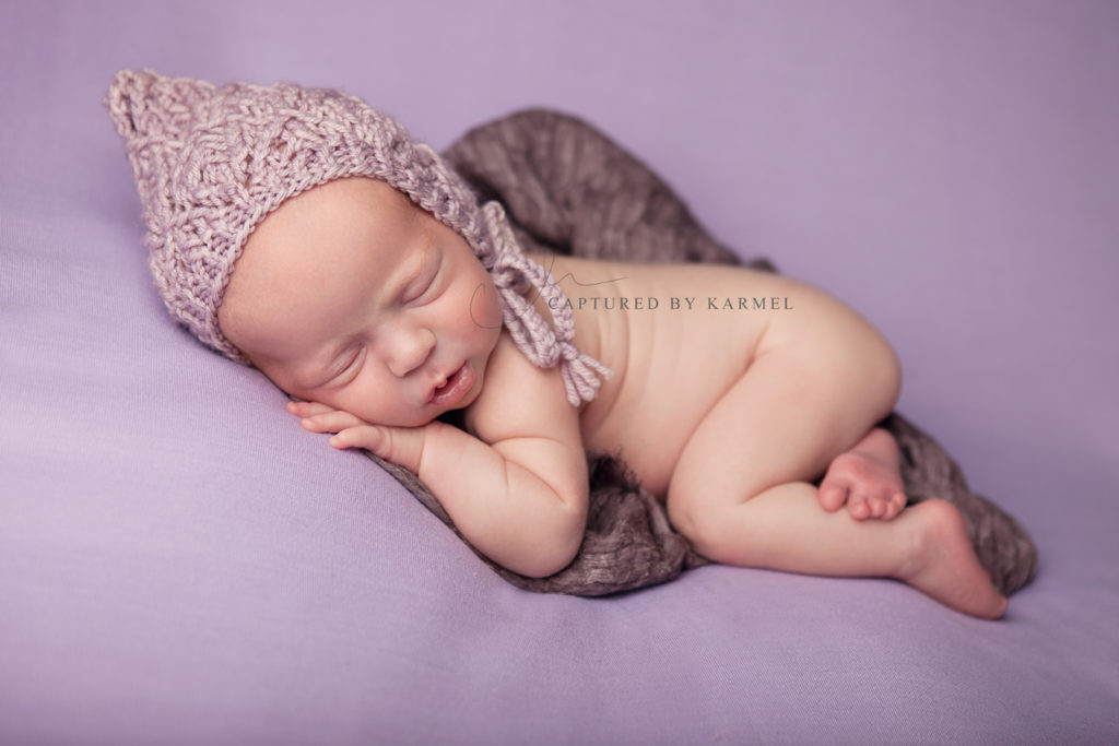 newborn sleeping on purple backdrop and wearing knitted hat prop